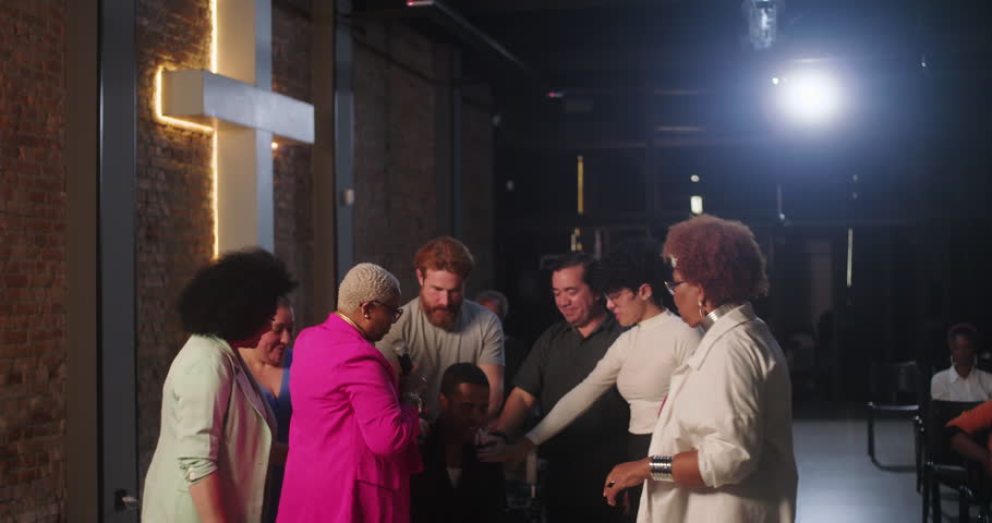Diverse group of people offering support and prayer, hands on a man during emotional moment, spiritual gathering with cross in the background