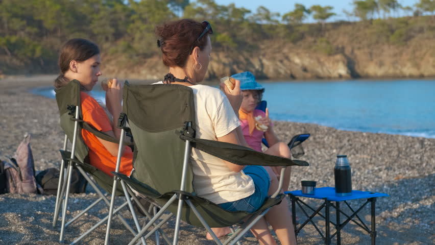Family enjoying snacks on beach chairs by the sea. Family members resting on folding chairs at pebble shoreline, sharing snacks while overlooking serene ocean landscape during summer leisure moment