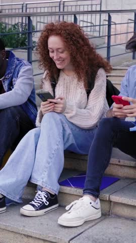 Three multiracial friends gathering outside university, sitting on staircases using phone together, laughing and having a fun talk. Vertical footage with copy space.
