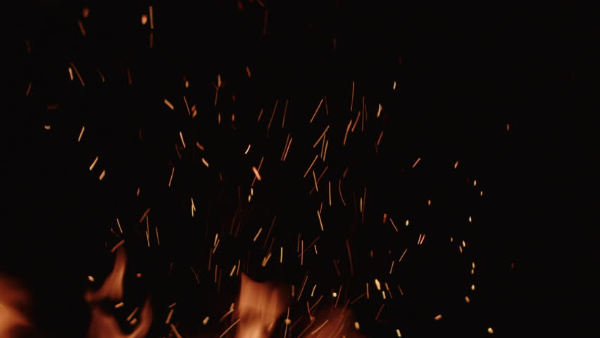 A dynamic close-up of a roaring fire showcases vibrant flames dancing against a dark backdrop, sending a shower of golden sparks soaring upwards