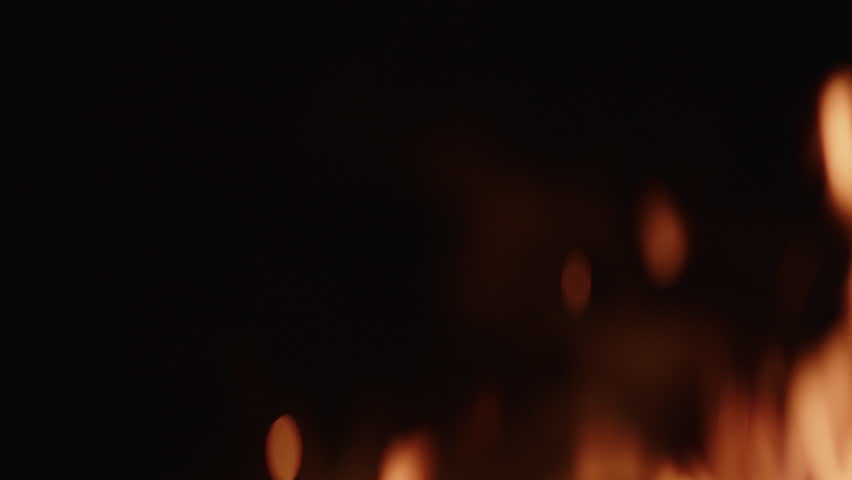 A close-up view of a flickering orange-red fire shows flames dancing and casting a warm, intense glow against a dark background as it burns brightly