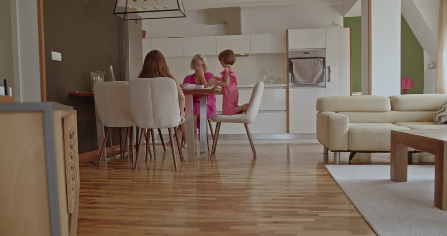 A mother and her two daughters are sitting at the dining table and interacting. The mother is wearing a pink dress and the daughters are wearing pink pajamas.