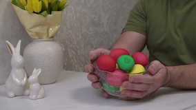 4k. A man is twirling a glass bowl with Easter eggs painted with food coloring in different colors, sitting at a table with a bouquet of yellow tulips and bunny figurines. - Powered by Shutterstock - Get 15% off with code: PIKWIZARD15