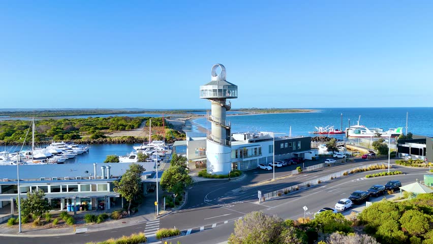 Aerial footage of Queenscliff Marina with lighthouse, boats, and coastline under clear blue skies