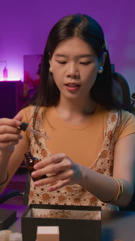 Vertical medium shot of beautiful young Chinese female influencer sitting in studio, advertising merchandise to subscribers on livestream, taking face serum out of box and applying on skin to test
