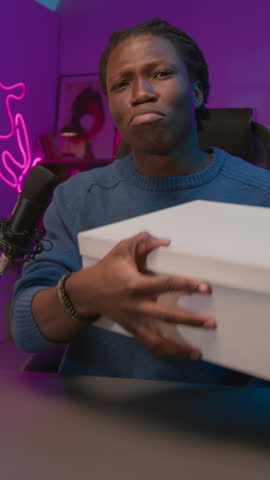 Vertical medium UGC shot of happy, vivacious black male blogger opening box with sneakers during live show, checking sole, praising trendy design, while looking at camera