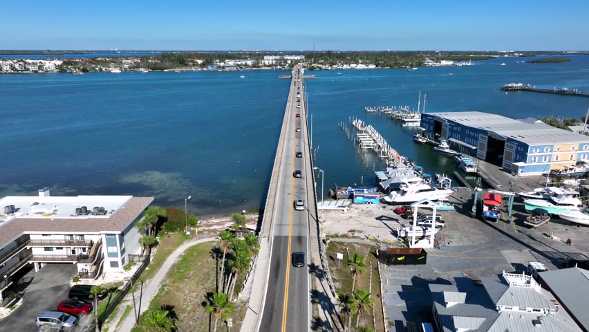 Aerial birds eye shot of cars on suspension bridge Crossing Manatee River in Brandeton, Florida. Sunny day with blue sky. Port and pier with docking sailing boats. Island on other side. Summer season