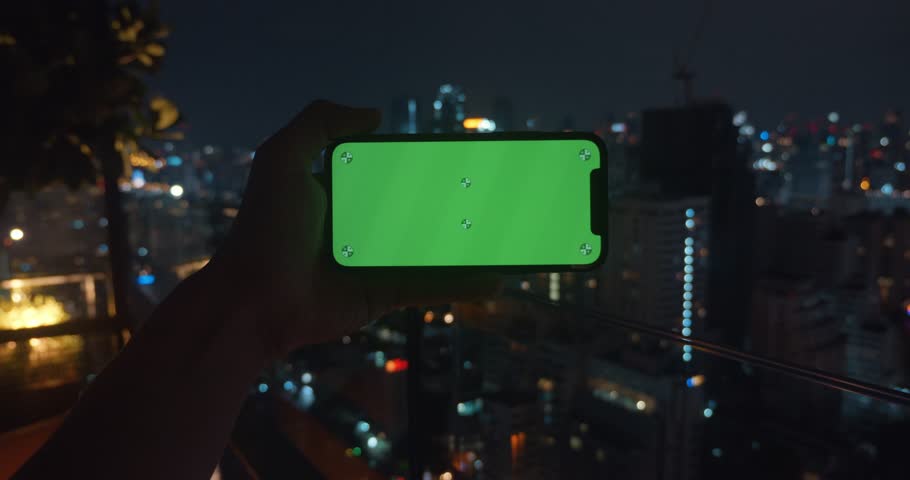 Close-up of hand holding phone horizontally with city illuminated by lights at night. View of evening skyscrapers city through gadget screen. Smartphone with chromakey in night city for design mockup.