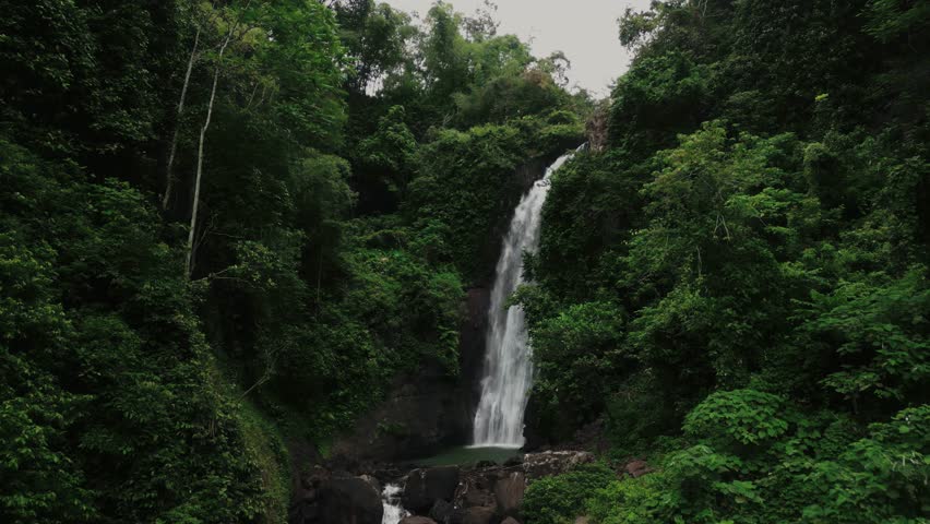 Aerial drone shot of Hidden waterfall in tropical rainforest jungle, drone flying towards the waterfall and slowly tilting down the camera, revealing some huge rocks