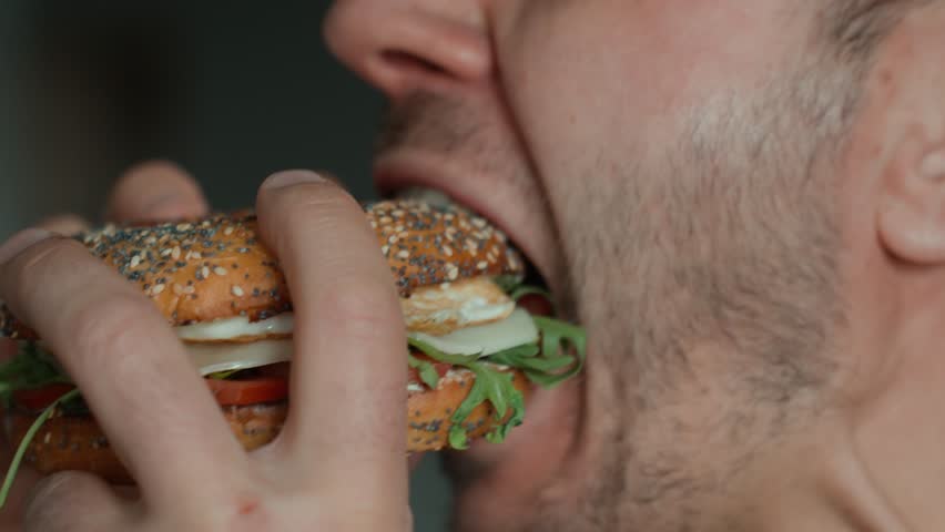 Man takes big bite of seeded bagel filled with egg, cheese, tomato and greens. Hungry man eats sandwich at kitchen, close up