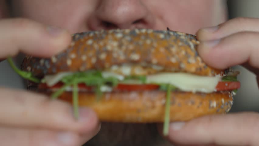 Close up of mouth takes big bite of seeded bagel filled with egg, cheese, tomato and greens. Hungry man eats sandwich