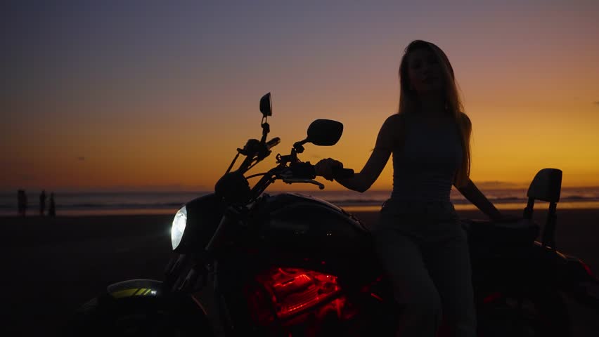 Cool girl posing with motorcycle at sunset, woman with bike on the beach in dark setting. Feeling freedom and independence, having moto trips and exciting adventures.