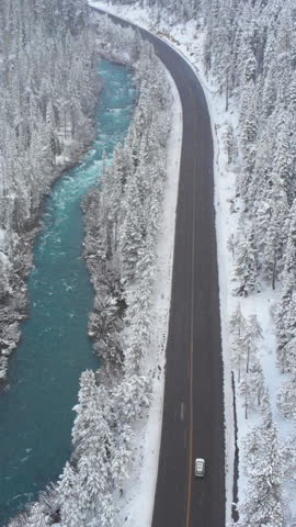 AERIAL: White car drives down empty road running along the beautiful emerald river in the cold Californian winter. Tourists on road trip cruising through the idyllic snow covered countryside and woods
