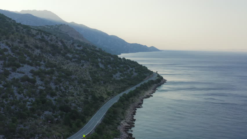 Aerial wide shot of winding coastal road, hills and sea at twilight. Mountains fading into the horizon. Gentle gusts visible on tranquil sea. Jadranska Magistrala, Croatia.