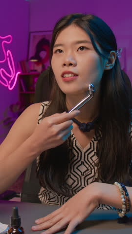Vertical medium shot of young Chinese female lifestyle blogger promoting new model of face massage roller to social media audience on live stream, with usage tutorial