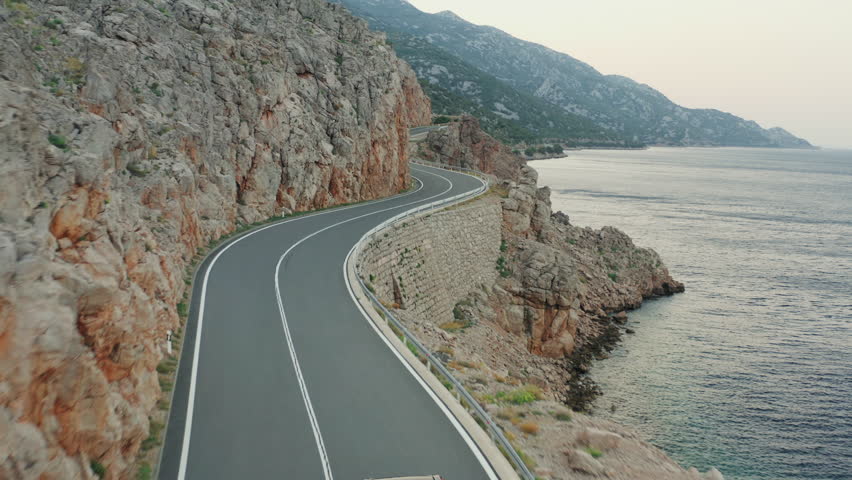 Aerial push in above winding coastal road carved into rocky cliffside at twilight. Calm sea on the right. Jadranska Magistrala, Croatia