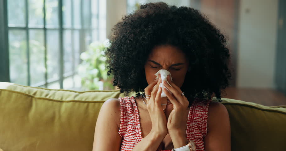 Woman, sneeze and tissue on sofa in house, sick and sinusitis with flu, allergy and rest in living room. African person, cleaning nose and nasal inflammation with cold, mucus or hygiene at apartment