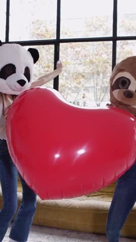 Vertical HD video of two cheerful women wearing panda and sloth masks dancing while holding a big inflatable red heart and raising arms in a modern apartment
