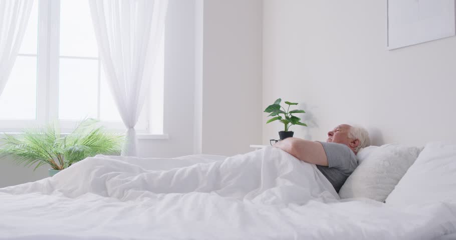 Senior elderly man wearing sleepwear waking up in bed and stretching his arms in bedroom at home. Gray haired retired male person doing morning exercises for health indoors. 4k video.