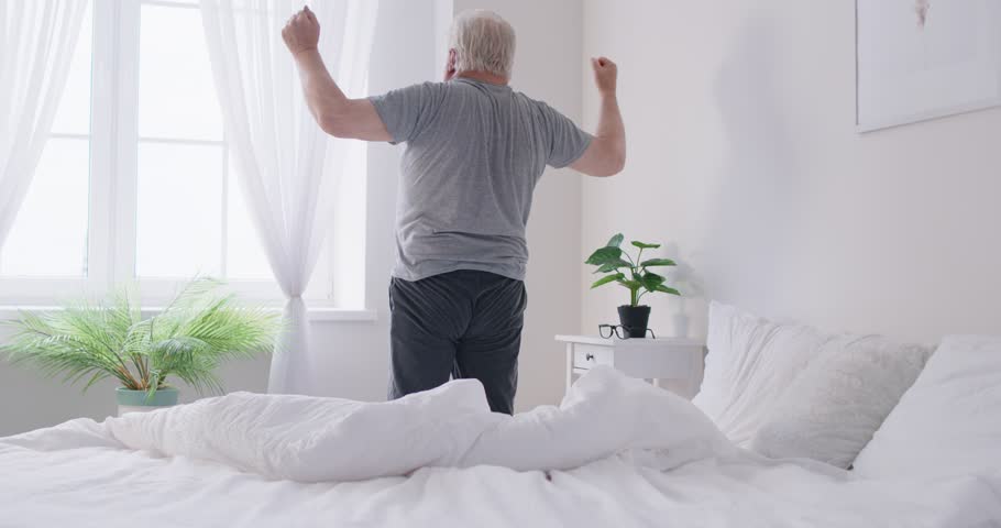 Senior elderly man wearing sleepwear waking up in bed and stretching his arms in bedroom at home. Gray haired retired male person doing morning exercises for health indoors. 4k video.