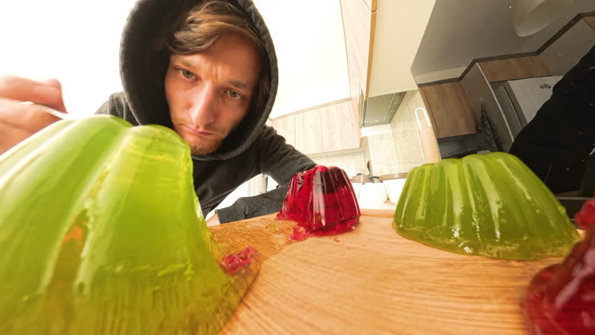 A man places a tray with red and green homemade jelly back into the refrigerator while eating a piece directly from it with a spoon. Wide-angle view from inside the fridge