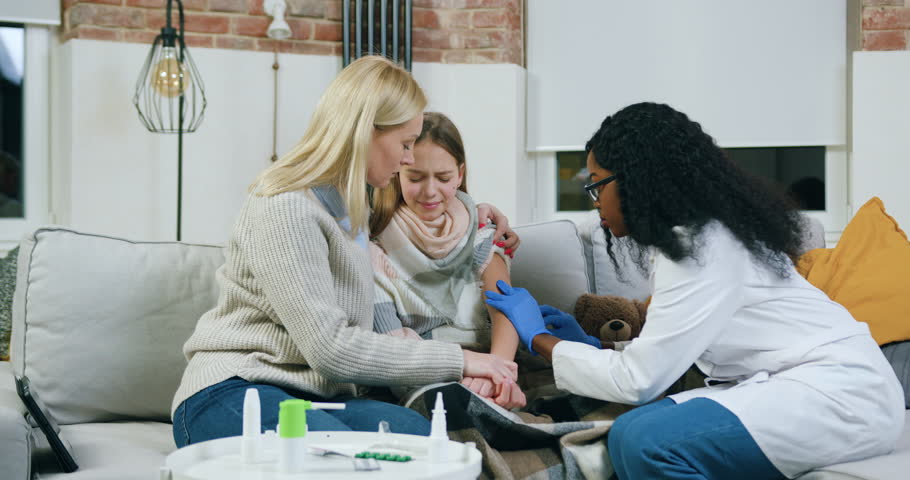 Attractive positive high-skilled black-skinned private female doctor giving an injection to unhealthy teen girl which coucht cold and recieves treatment at home under mother's supervision