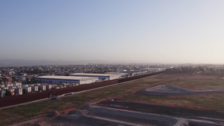 Aerial view of the U.S.-Mexico border wall with urban development on one side and open land on the other at sunset.