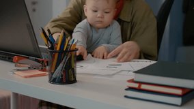Young mother attempts to work on her laptop with a baby cradled in her arms, sorting through various notes and papers in a cluttered home office setting. - Powered by Shutterstock - Get 15% off with code: PIKWIZARD15