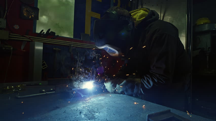 Metal worker in safety work gear welding steel in a dark manufacturing workshop, medium shot. Home business and small company concepts.