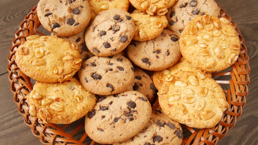 Homemade chocolate chip and peanut cookies gently rotating in rustic wicker basket, showcasing golden-brown color, delectable texture, and irresistible bakery-fresh appeal.