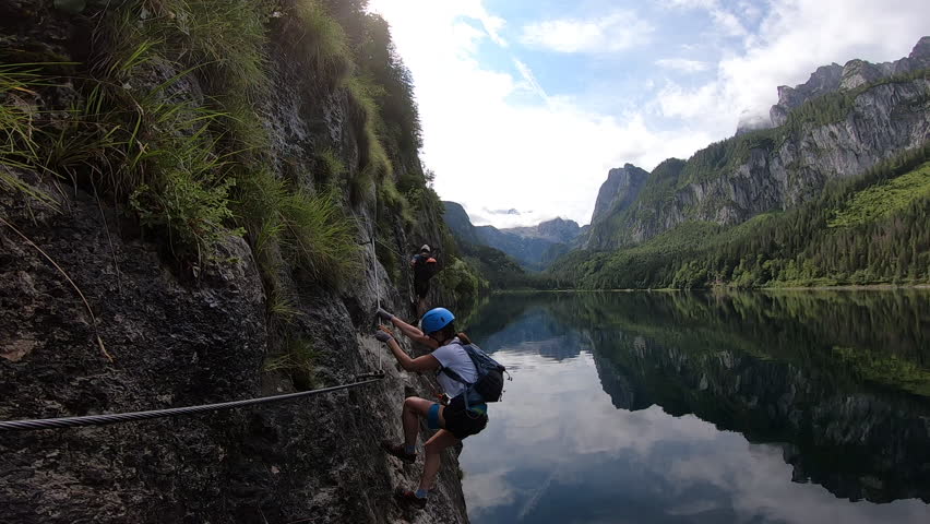 A climber carefully ascends a steep via ferrata, enjoying stunning lake reflections and mountain scenery.