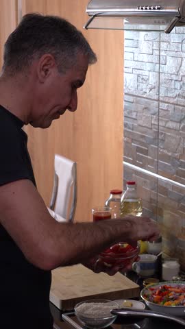 Handsome Middle-Aged Man Adds Cherry Tomatoes to a Chicken and Vegetable Dish While Cooking on Induction Cooktop - Panning Shot. Lifestyle of middle-aged men. Healthy eating. Home Cooking.