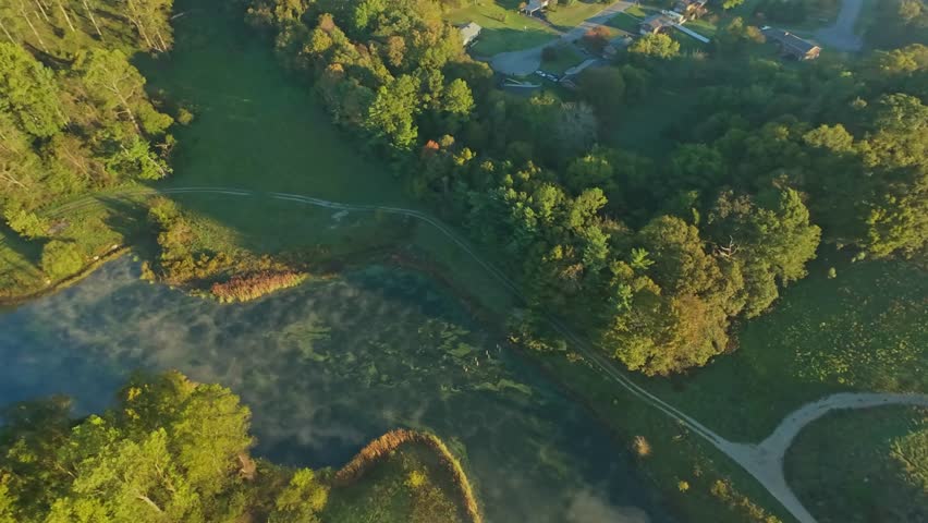 Drone image of misty pond surrounded by trees, grassy fields, and winding paths in serene suburban park during sunrise. Aerial View of Green Parkland and Pond in Morning Light
