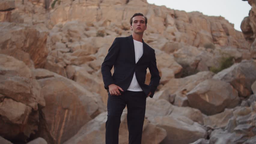 Handsome guy in a stylish suit poses in the mountains. Male fashion model in a blue business suit poses standing on big stones in rocks. Handsome young guy model posing for camera in the mountains. 