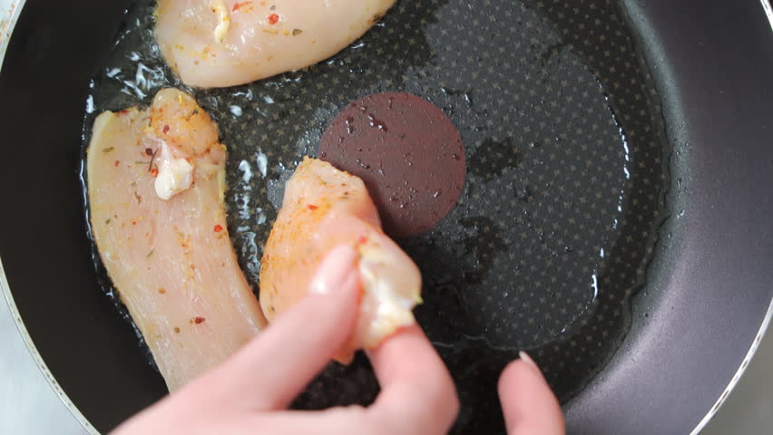 Raw chicken breasts seasoned with spices placed into a hot non-stick frying pan with oil, overhead view captures sizzling start of the cooking process