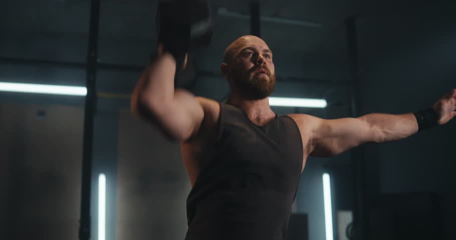 Focused male athlete in a gray tank top lifting a heavy dumbbell while doing sports in a modern gym