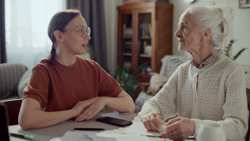 Young woman smiling and talking with hand on shoulder of elderly grandmother, sharing support during paperwork at home, both sitting at table with bills and documents