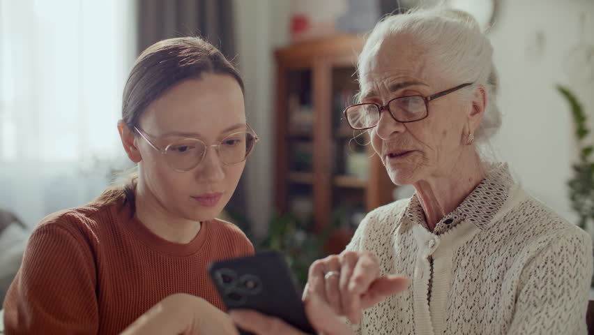 Young woman teaching elderly grandmother how to use smartphone, helping her to explore modern technology at home