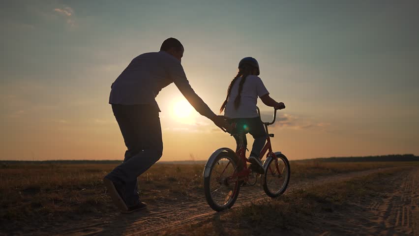 Father teaches child to ride bike at sunset. Child learns to balance on bike with father support. Happy family moment outdoors. Father guiding child on dirt road. Bonding over bike riding lessons.