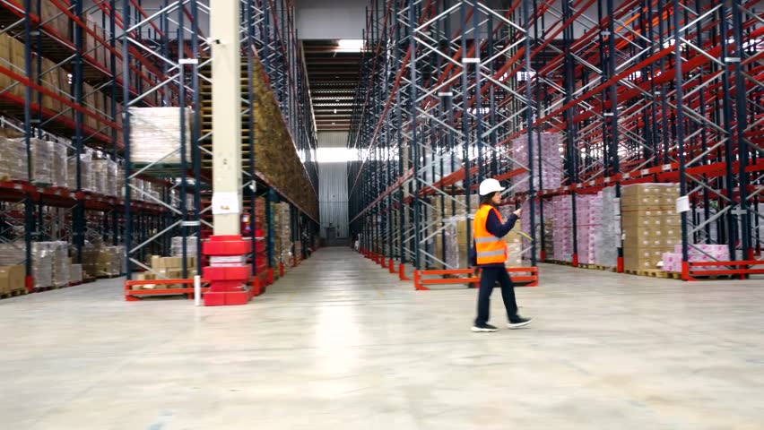 Female supervisor wearing safety vest and helmet inspecting goods and merchandise on shelves in large distribution center