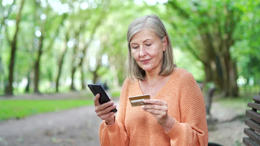 Mature woman sitting on bench outdoors using smartphone and credit card. She is smiling and engaged in digital banking or online shopping in tranquil park setting. Modern lifestyle and technology