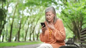 Elderly female with gray hair sitting on park bench looking at phone with concern, receiving bad news outdoors. Mature woman wearing orange sweater looking at smartphone with distressed expression - Powered by Shutterstock - Get 15% off with code: PIKWIZARD15
