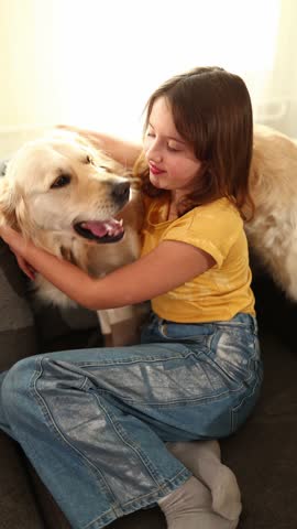 A young girl with brown hair smiles as she hugs a golden retriever standing on a couch. The well-lit living room adds warmth to their joyful moment together.