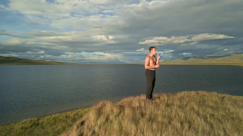 A man stands on a hill above a mountain lake and cheerfully blows soap bubbles at sunset, during the golden hour.