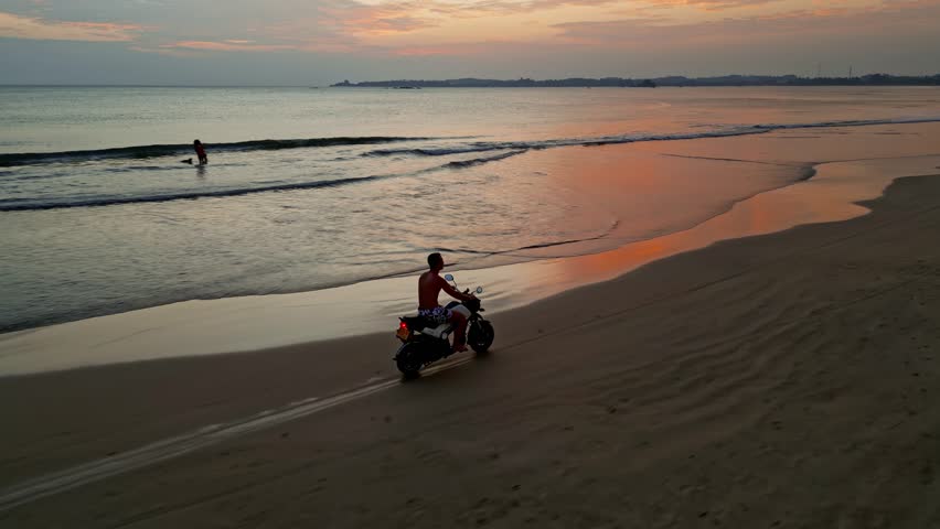 Man driving moto along ocean beach at sunset. Aerial view back side
