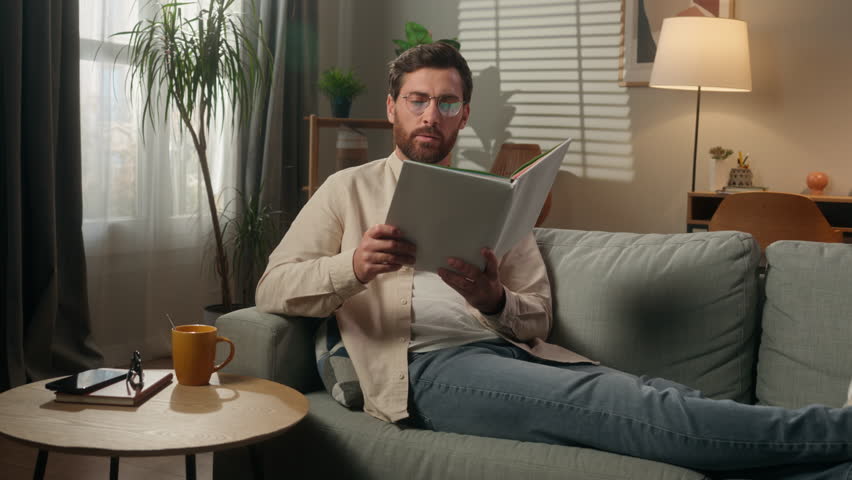 Caucasian man reading book alone at home male homeowner reader smart guy businessman in eyeglasses read literature on couch sofa paper pages textbook hobby leisure rest in apartment studying knowledge