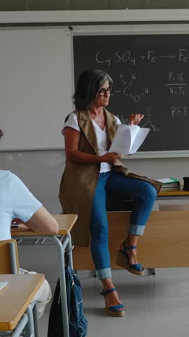 Vertical HD video of mature female teacher explaining exam or educational exercises to her high school students at classroom. Back to school and education lifestyle concept.