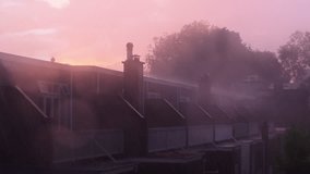 A row of houses with chimneys is seen under a pink-hued sky during rainfall. Raindrops blur the view, and trees in the background add depth to the misty atmosphere. The sunset casts a soft glow. - Powered by Shutterstock - Get 15% off with code: PIKWIZARD15