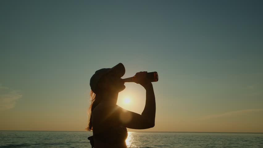 Silhouette of a young long haired woman drinking from a bottle by the sea at sunset. A refreshing sip of beer on a hot summer evening on vacation.
