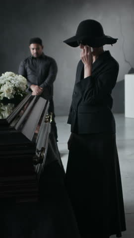 Vertical shot of grieving woman in black, large hat and sunglasses, holding tissue, standing near coffin adorned with white flowers at funeral